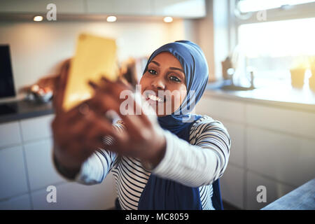 Souriante jeune femme arabe portant un hijab en tenant vos autoportraits avec son portable alors qu'il était assis dans sa cuisine à la maison Banque D'Images