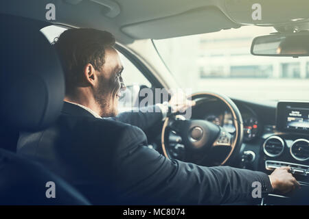 Young businessman smiling et changement des stations radio sur sa voiture en conduisant à travers la ville pendant son trajet du matin au travail Banque D'Images