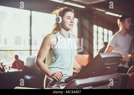 Thème est le sport et la musique. Une belle femme gonflé s'exécute dans la salle de sport sur un tapis roulant. Sur sa tête un casque blanc sont grandes, la jeune fille à l'écoute de la musique pendant une séance de cardio pour la perte de poids Banque D'Images