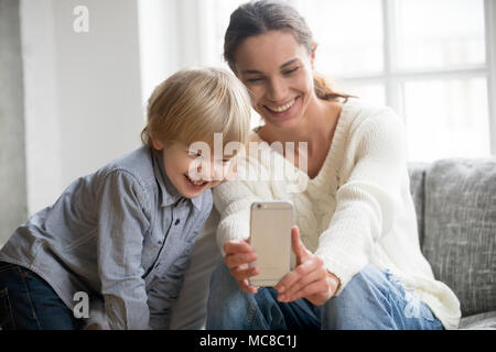 Heureuse fête de rire en tenant avec petit-fils selfies sur smartphone à la maison, maman simple sourire et mignon adopté Garçon jouant en posant pour la photo sel Banque D'Images