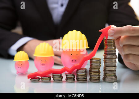 Entrepreneur's Hand Holding flèche au-dessus de pièces empilées montrant vers le haut en face de Piggy Bank Banque D'Images