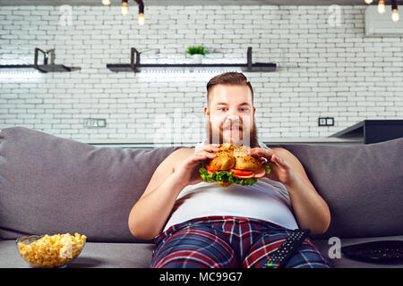 L'homme drôle Fat en pyjama de manger un hamburger assis sur le canapé à la maison. Banque D'Images