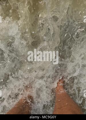 Vue de dessus de la jambe d'un homme debout sur le sable et baigné par les vagues de l'océan. Banque D'Images