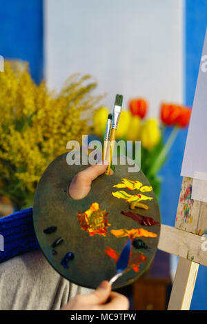 Artiste en pull bleu attire les peintures à l'huile se mélange avec couteau close-up. Il est titulaire en poils de pinceau et de la palette. Fleurs dans l'arrière-plan Banque D'Images