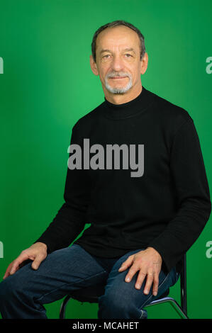 Bel homme mature aux cheveux gris et avait la barbe en noir pull et jeans foncé assis sur la chaise sur un fond vert en Studio Banque D'Images