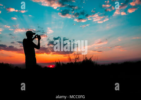 Sombre Silhouette de jeune homme photographier le coucher du soleil sur l'appareil photo du smartphone et de l'arrière-plan coloré du ciel du soir. Soleil au lever du soleil, dans les produits de Banque D'Images