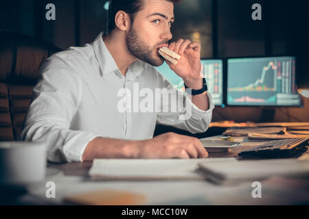 Homme d'affaires occupé ayant déjeuner tardif eating sandwich dans office travaillant de nuit, agent de négociation de titres en ligne et de manger à la collation à l'ordinateur s'workp Banque D'Images