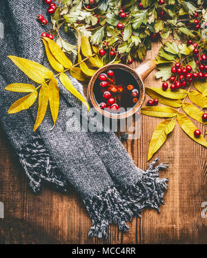 Tasse de thé de fines herbes avec des baies d'aubépine d'automne , feuilles d'automne et foulard sur fond de bois rustique, vue du dessus. Banque D'Images