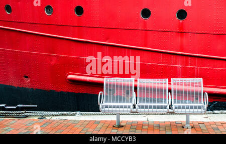 3 chaises en métal blanc vide sur une allée en brique en face d'un grand rouge lumineux coque de bateau avec hublots à Baltimore's Inner Harbor Banque D'Images