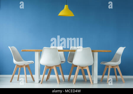 Bâtiment moderne, blanc et des chaises en bois autour d'une table à manger et lampe jaune contre le mur à l'arrière-plan bleu dans un intérieur de style minimal Banque D'Images