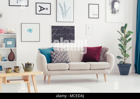 Table en bois près de settee avec coussins dans un salon intérieur avec des ficus et des affiches Banque D'Images