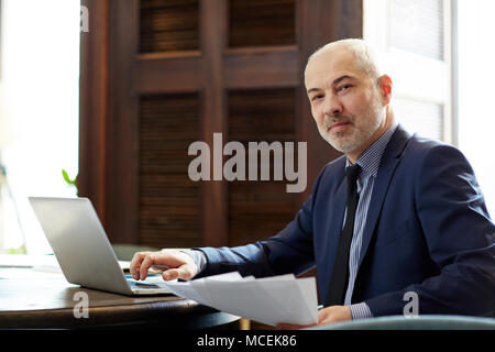 Portrait of smiling Caucasian businessman aux cheveux gris assis à un ordinateur portable avec documents et looking at camera Banque D'Images