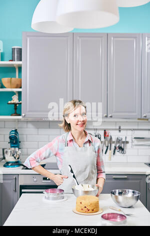Young attractive Caucasian woman en tablier, debout dans la cuisine avec du pastry and smiling at camera Banque D'Images