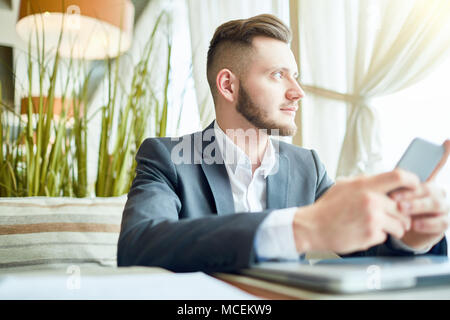 Les jeunes barbus attractive businessman sitting at cafe table avec le smartphone et en regardant par la fenêtre pensivement Banque D'Images