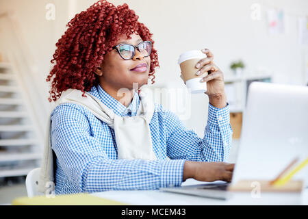 Portrait of attractive African American Woman with red cheveux bouclés assis à un ordinateur portable avec du café à emporter et sourire rêveur Banque D'Images