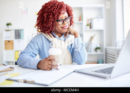 Female office worker noir élégant avec les cheveux bouclés rouge assis à un bureau avec du café à emporter, la lecture des nouvelles sur laptop and smiling Banque D'Images
