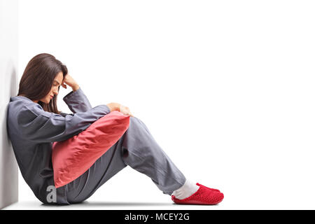 Triste jeune femme en pyjama assis sur le plancher et la tenue d'un oreiller isolé sur fond blanc Banque D'Images