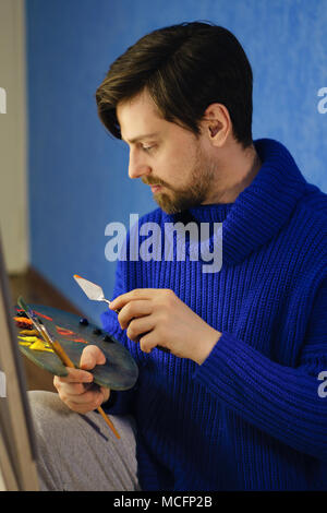 Artiste en pull bleu attire l'allie de la peinture à l'huile avec couteau à palette. Il est titulaire en poils de pinceau et de la palette. Banque D'Images