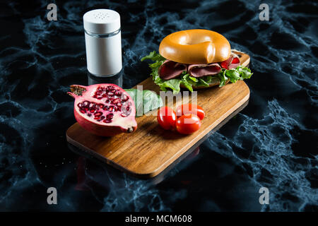 Bagel jambon sur une planche à découper en bois avec grenade et tomates cerises. Banque D'Images