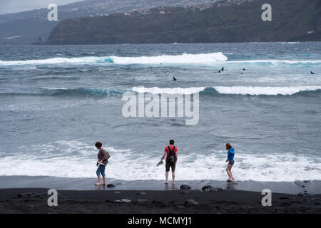 Puerto de la Cruz, Tenerife, Canaries ; touristes tester la température de l'eau sur une plage de sable noir alors que les surfeurs pièges pour prendre les vagues. Banque D'Images