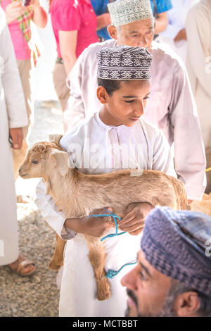 Nizwa, Oman - Mar 23, 2018 : Jeune garçon omanais dans une tenue traditionnelle omanaise blanc tenant un bébé chèvre. Banque D'Images