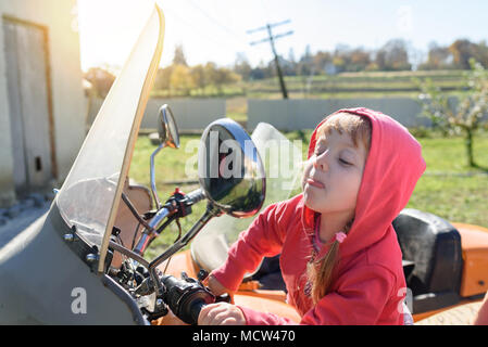 Funny little girl est assis sur la moto, regarde dans le miroir et montre de la langue, de l'enfance insouciante Banque D'Images