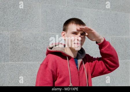 Le gars dans la capuche rouge et se tient au mur et couvre ses yeux avec sa main de la lumière du soleil. Banque D'Images