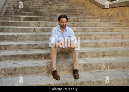 Young attractive l'homme assis sur le travail avec des escaliers de tablette numérique pad à la recherche satisfait et confiant dans les nouvelles technologies d'affaires et de li Banque D'Images