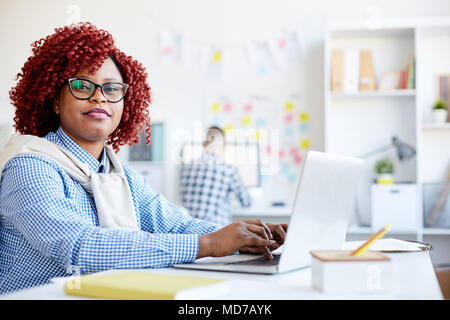 Portrait of female office worker noir élégant avec les cheveux bouclés rouge looking at camera while sitting at desk et working on laptop Banque D'Images