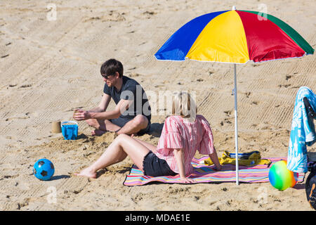 Bournemouth, Dorset, UK. 19 avril 2018. Météo France : belle chaude journée ensoleillée à plages de Bournemouth avec un ciel bleu et soleil ininterrompue, en tant que visiteurs, chef de la mer pour profiter de la journée la plus chaude de l'année jusqu'à présent. Couple sur la plage, sous un parasol aux couleurs vives. Credit : Carolyn Jenkins/Alamy Live News Banque D'Images