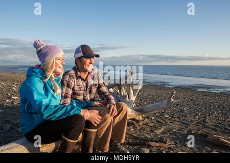 Un jeune couple est assis sur un morceau de bois flotté sur une plage donnant sur l'océan au coucher du soleil ; Anchorage, Alaska, États-Unis d'Amérique Banque D'Images