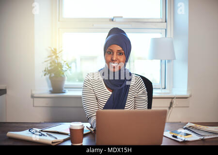 Jeune femme entrepreneur arabe portant un hijab avec confiance sourire tout en travaillant sur un ordinateur portable dans son bureau à domicile Banque D'Images