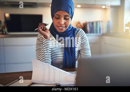 Jeune femme portant un hijab en arabe à travers la lecture de documents et de travailler sur un ordinateur portable tout en étant assis à une table dans sa cuisine Banque D'Images