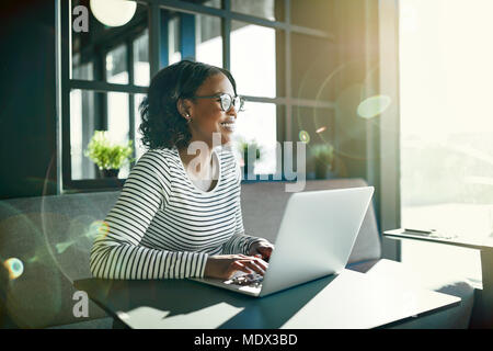 Souriante jeune femme africaine portant des lunettes à la recherche par la fenêtre tout en étant assis à une table en ligne de travail avec un ordinateur portable Banque D'Images