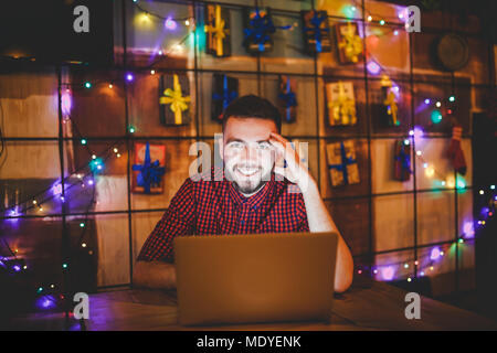 Un beau jeune homme de race blanche avec barbe et sourire à pleines dents dans une chemise à carreaux rouge fonctionne derrière un ordinateur portable gris assis à une table en bois. Mains o Banque D'Images