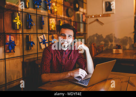 Un beau jeune homme de race blanche avec barbe et sourire à pleines dents en chemise à carreaux rouge fonctionne derrière laptop sitting at table en bois. Utilise mobile détient Banque D'Images