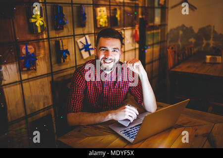 Un beau jeune homme de race blanche avec barbe et sourire à pleines dents en chemise à carreaux rouge fonctionne derrière laptop sitting at table en bois. Utilise mobile détient Banque D'Images