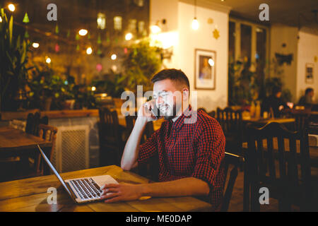 Beau jeune homme de race blanche avec barbe et sourire à pleines dents en chemise rouge fonctionne en arrière, les mains sur le clavier d'ordinateur portable assis à table en bois. Utilise des appels sur mo Banque D'Images