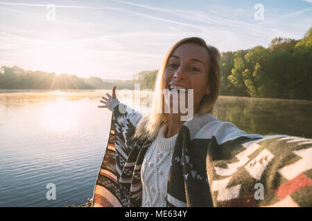 Jeune femme par le lac au lever du soleil prend un portrait selfies en utilisant un téléphone mobile, belle réflexion sur la surface de l'eau, modèle portant une couverture. Billet d Banque D'Images