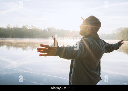 Jeune homme bras tendus par le lac au lever du soleil, jouissant de la liberté et de la vie, les gens voyagent bien-être concept. Tourné en France, en Europe. Banque D'Images