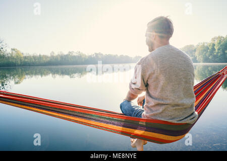 Jeune homme sur hamac détente au bord de la lac dans le matin, la lumière du soleil. Les gens voyagent bien-être la paresse concept. France, Europe Banque D'Images