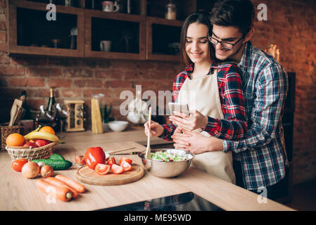 L'amour heureux couple prépare un dîner romantique sur la cuisine. Smiling mari épouse sa femme en tablier qui est en train de préparer une salade de légumes Banque D'Images