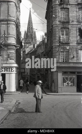 1950, historique, un gentlemam avec un bâton de marche traverse une route dans le centre-ville de la ville de Caen, France. Une flèche de l'Abbaye aux Hommes peut être voir dans la distance. Banque D'Images