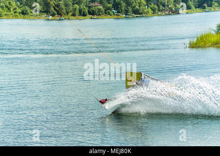 Wakeboarder saute d'un tremplin derrière une corde et fait un vague sur l'eau. Banque D'Images