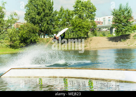 Wakeboarder saute d'un tremplin derrière une corde et fait un vague sur l'eau. Banque D'Images