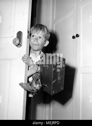 Un jeune garçon vient prudemment dans la maison avec ses vêtements sales, les chaussures boueuses, et sa boîte à lunch après l'école, ca. 1964. Banque D'Images