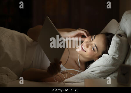 Happy girl portant des écouteurs à regarder des vidéos en ligne dans un comprimé allongé sur un lit dans la nuit à la maison Banque D'Images