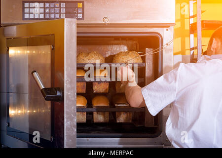 Un mâle Baker en uniforme blanc et un tablier beige cuit le pain d'un four industriel. Travailler à la boulangerie Banque D'Images