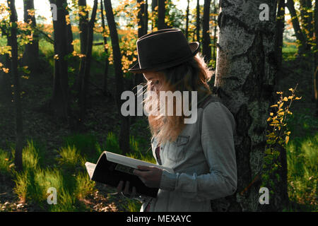 Young caucasian woman artiste portant un chapeau élégant, debout dans une forêt de bouleaux tenant son portable Banque D'Images