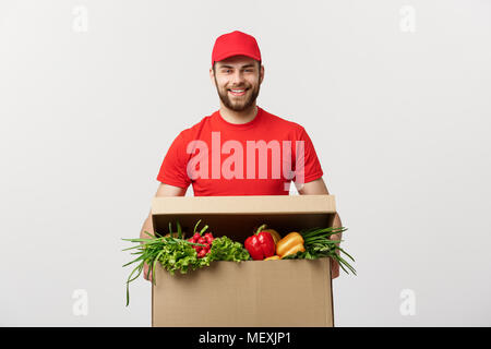 Concept de livraison : Handsome Young man in courrier de livraison d'épicerie uniforme rouge avec boîte d'épicerie avec des fruits et légumes Banque D'Images